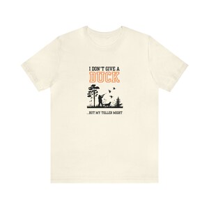 Op de afbeelding: Een wit T-shirt met een zwarte en oranje grafische print. De afbeelding toont een jager met een hond en vliegende eenden. De tekst luidt "Ik geef geen moer...maar mijn toller misschien."