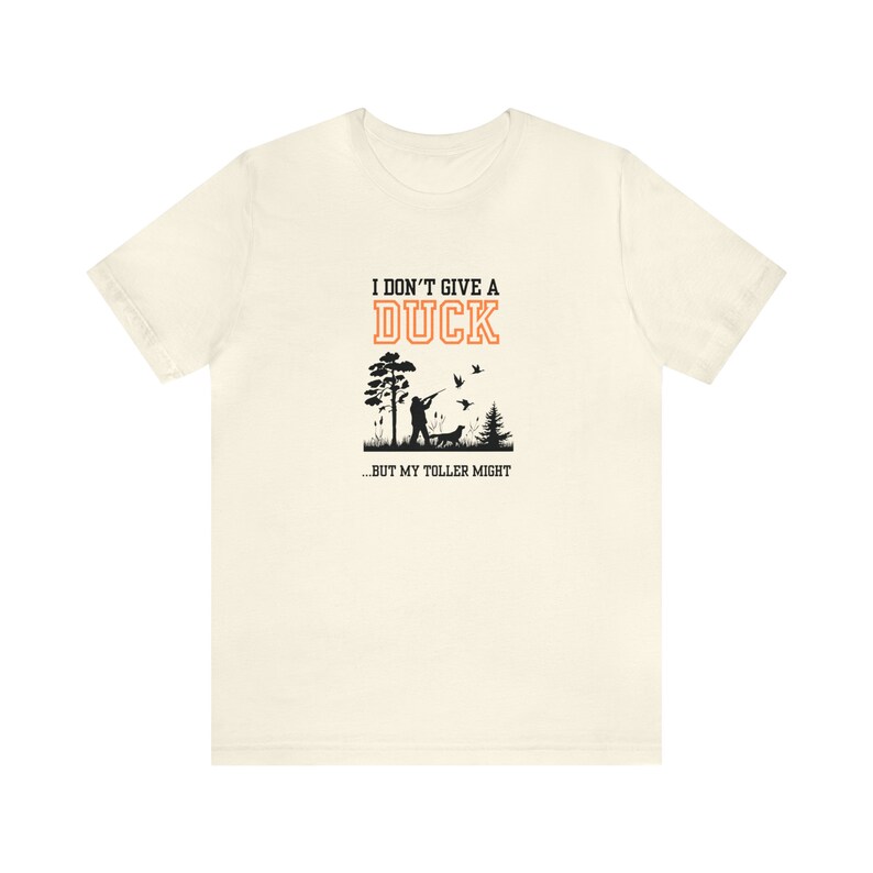 Op de afbeelding: Een wit T-shirt met een zwarte en oranje grafische print. De afbeelding toont een jager met een hond en vliegende eenden. De tekst luidt "Ik geef geen moer...maar mijn toller misschien."
