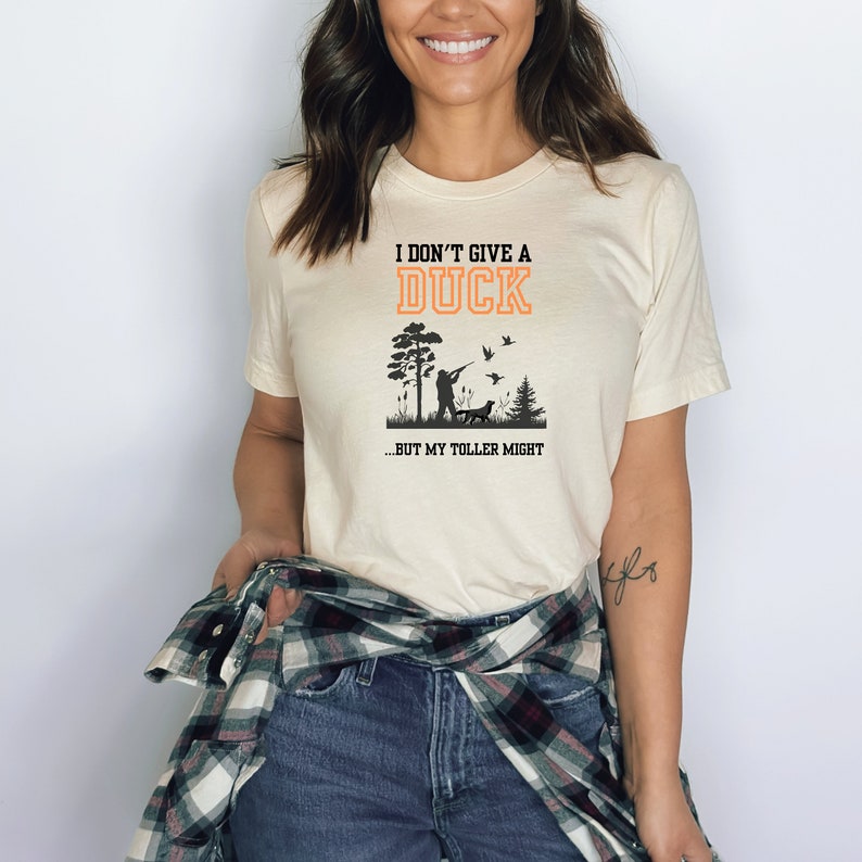 Op de afbeelding: Een wit T-shirt met de tekst "I don't give a duck...but my toller might" in oranje en zwart. De afbeelding toont een silhouet van een persoon die eenden jaagt met een hond.