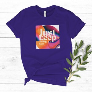 May include: Purple t-shirt with a graphic design of a colorful abstract shape with the text "Just keep circling back" in a retro font.