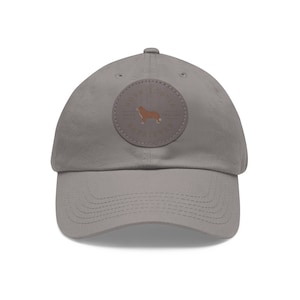 Peut inclure: Une casquette de baseball grise avec un patch en cuir brun représentant la silhouette d'un Nova Scotia Duck Tolling Retriever et le texte "Nova Scotia Duck Tolling Retriever Since 1998"