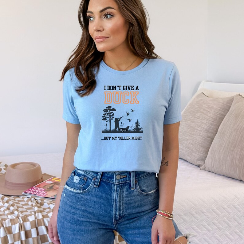 Op de afbeelding: Een lichtblauw t-shirt met een afbeelding van een persoon die eenden jaagt met een hond. De tekst op het shirt luidt "I don't give a duck...but my toller might."
