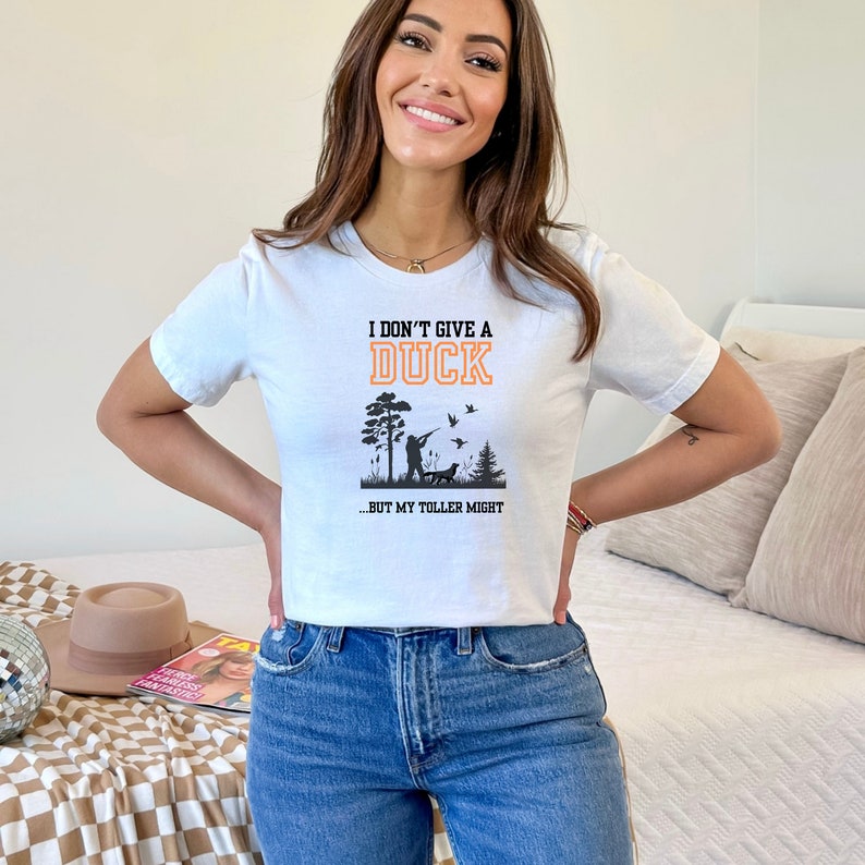 Op de afbeelding: Wit T-shirt met een zwarte en oranje afbeelding met de tekst "Ik geef geen moer om een eend...maar mijn toller misschien". De afbeelding toont een silhouet van een persoon die eenden jaagt met een hond.