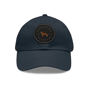 Peut inclure: Une casquette de baseball bleu marine avec un patch en cuir marron représentant la silhouette d'un chien et le texte "Nova Scotia Duck Tolling Retriever".