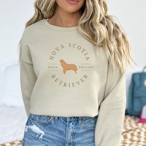 May include: A beige sweatshirt with a brown graphic of a Nova Scotia Duck Tolling Retriever dog. The graphic includes the text "Nova Scotia Duck Tolling Retriever".