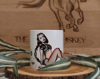 May include: White ceramic mug with a colorful illustration of a woman with a scorpion tail and stars. The mug is on a wooden coaster with green leaves.