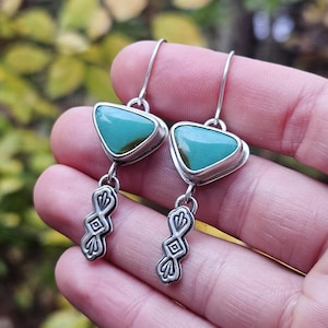 May include: A pair of silver earrings with turquoise triangular stones. The earrings have a decorative silver dangle with an etched design. The earrings are displayed against a blurred background of green and yellow foliage.