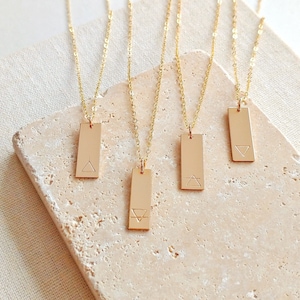 May include: Four gold necklaces with rectangular pendants featuring different geometric symbols. The pendants are engraved with triangles and a circle.