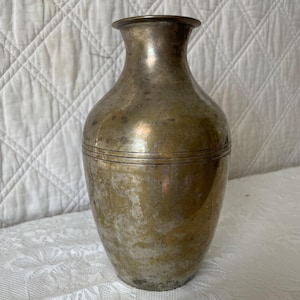 May include: A brass vase with a tapered neck and a rounded body. The metal surface features a weathered, tarnished patina with visible variations in color and texture. Two thin, horizontal engraved lines circle the midsection of the vase.