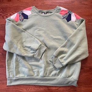 May include: Sage green sweatshirt with floral and geometric patchwork detail on the shoulders. The long-sleeved top has ribbed cuffs and hem. The patchwork includes pink, white, and navy blue fabric.