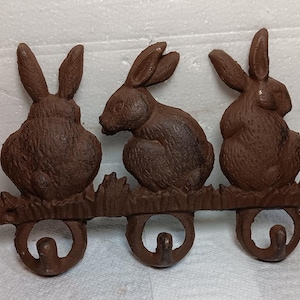 May include: A cast iron wall mounted coat rack with three hooks shaped like rabbits. The rabbits are brown and have a rustic finish. The hooks are attached to a wooden base with a grass design.
