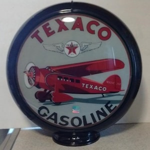 Könnte beinhalten: Eine Vintage-Texaco-Tankstellenkugel mit einem roten Flugzeug, das über einem weißen Stern mit Flügeln fliegt. Die Kugel ist schwarz mit weißem Hintergrund und den Worten "Texaco Gasoline" in schwarzer Schrift.