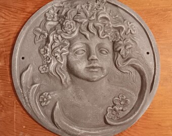 Può includere: Una placca da parete rotonda e decorativa di colore grigio con un'immagine scolpita di un viso di donna con fiori nei capelli.