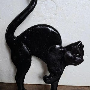 May include: A black cast iron figurine of a cat in an arched-back posture with its tail held high. The cat is depicted with its head lowered and one front paw slightly raised, standing on a rectangular base with a textured, matte finish.