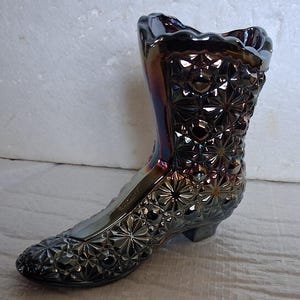 May include: An iridescent glass boot-shaped vase. The vase has a textured surface with a starburst pattern. The boot is dark with purple and gold hues, and has a scalloped top.
