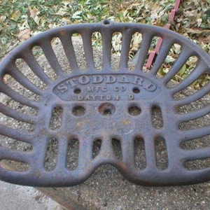 Puede incluir: Un asiento de tractor vintage de hierro fundido con un acabado negro y las palabras "Stoddard Mfg Co Dayton O" en relieve en la parte delantera.
