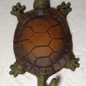 May include: A brown and green ceramic turtle figurine with a textured shell. The turtle has a long tail that curls up and is used as a hook.