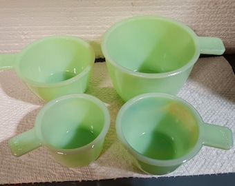 Puede incluir: Cuatro tazas medidoras verde jade de diferentes tamaños dispuestas sobre una toalla de papel blanca. Cada taza tiene un asa. Las tazas son de vidrio translúcido verde claro.