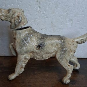 May include: A vintage cast iron dog figurine with a detached head piece. The dog is painted in an off-white color with dark gray and brown weathered patches throughout its body. It stands in a pointing pose with one front leg raised.