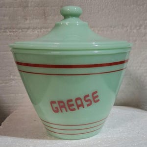 Puede incluir: Un tarro para grasa de vidrio jadeíta verde claro con tapa a juego. El tarro presenta rayas rojas horizontales en las secciones superior e inferior y la palabra GREASE impresa en letras rojas en el frente del cuerpo cónico.