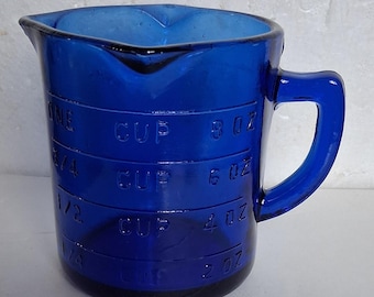 May include: A cobalt blue glass measuring cup with a handle and a pouring spout. The exterior features embossed measurement markings in cups and ounces, including 1/4, 1/2, 3/4, and one cup, as well as 2 oz, 4 oz, 6 oz, and 8 oz increments.