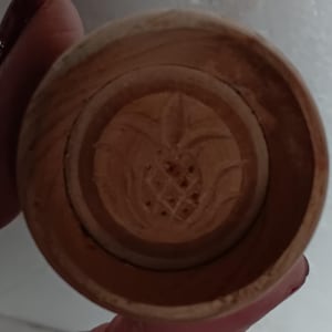 May include: A small, round, wooden object with a carved pineapple design in the center. The object is a light brown color and appears to be a stamp or mold. The pineapple design is detailed and intricate.