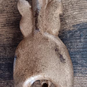 May include: A bronze-colored metal hook with a unique, abstract design. The hook has a curved top and a rounded base, with a textured surface. The hook is likely used for hanging items.