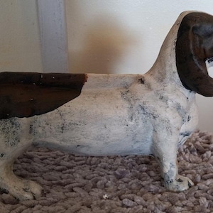 May include: A white and brown ceramic dog figurine with a distressed paint finish. The dog is standing on its four legs and has its head turned to the side. The dog is a basset hound breed.