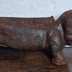 May include: A cast iron boot scraper shaped like a dachshund dog. The object features a dark, heavily rusted brown finish with a textured surface, a curled tail, and a flat top edge designed for cleaning footwear, showing visible screw hardware.