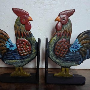 May include: Pair of cast iron rooster bookends. Each bookend features a detailed rooster design with red, blue, and yellow accents. The bookends are painted with a distressed finish, giving them a vintage look. The base is black.