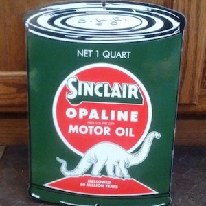 Puede incluir: Una señal vintage de lata de aceite de motor Sinclair Opaline. La señal es verde con un círculo rojo y blanco que presenta el logotipo de Sinclair y el texto "Opaline Motor Oil". Un dinosaurio blanco aparece en el círculo. El texto "Net 1 Quart" y "Mellowed 80 Million Years" también están en la señal.