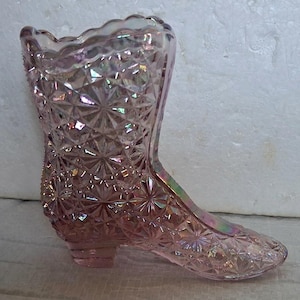 Op de afbeelding: Een roze glazen vaas in de vorm van een laars met een textuur van een sterrenpatroon. De vaas heeft een geschulpte bovenrand en een kleine hak. Het glas heeft een iriserende glans, die meerdere kleuren reflecteert. Een decoratief item.