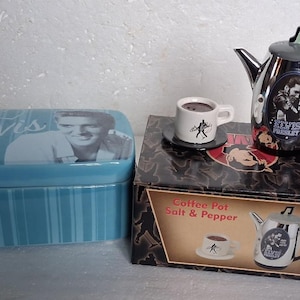 May include: A light blue ceramic box featuring Elvis Presley, a small white coffee cup with a black silhouette, and a silver and black coffee pot with an Elvis Presley image. The items are on a box that says "Coffee Pot Salt & Pepper."
