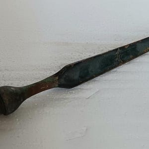 Puede incluir: Una antigua punta de lanza de bronce con una hoja larga y delgada y un mango cilíndrico. La pátina oscura y envejecida presenta toques de verde y marrón. Un artefacto histórico.