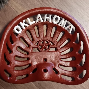 Puede incluir: Un asiento de tractor de hierro fundido rojo con la palabra "OKLAHOMA" en letras blancas en la parte superior. El asiento tiene un emblema de estrella en el centro y una serie de ranuras curvas para ventilación.