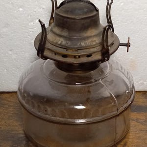 May include: A vintage glass oil lamp with a metal burner and a clear glass reservoir. The burner has a metal cap with the words "BEN  AN  NIP" inscribed on it.