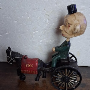 Könnte beinhalten: Vintage Gusseisenfigur eines Mannes, der eine Pferdekutsche fährt. Das Pferd ist schwarz mit einem roten Sattel, auf dem in goldener Schrift "THE NODDERS" steht. Der Fahrer trägt einen grünen Anzug und einen schwarzen Zylinder.