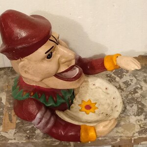 Op de afbeelding: Een rode, groene en gele gietijzeren spaarpot in de vorm van een nar met een grote mond en een bord in zijn hand.