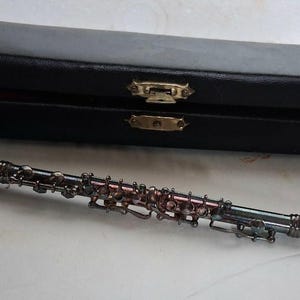 Puede incluir: Un clarinete negro con llaves plateadas y un estuche de cuero negro. El clarinete está sobre una superficie blanca junto a su estuche cerrado. El estuche tiene cierres dorados.