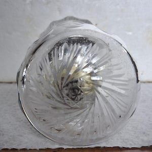 Puede incluir: Un jarrón de vidrio transparente con un patrón de remolino grabado en la parte inferior. El jarrón está vacío y tiene una base redonda y ancha.