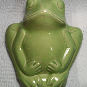 Puede incluir: Figura de rana de cerámica verde claro. La rana tiene ojos grandes y redondos, boca ancha y manos y pies detallados. La figura es un objeto decorativo, probablemente para el hogar o el jardín.