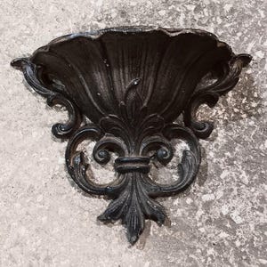 以下が含まれることがあります： 黒い装飾的な壁掛け棚。棚はスカラップのトップと、スクロールのディテールを備えた様式化された花のモチーフが特徴です。棚は金属製で、テクスチャード加工が施されています。