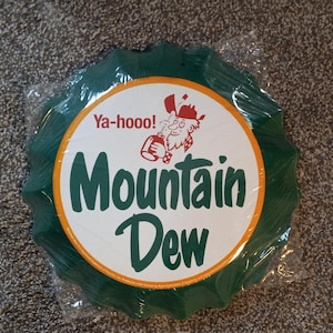 Puede incluir: Un tapón de botella de metal verde y blanco con las palabras "Ya-hoo! Mountain Dew" y una imagen de dibujos animados de un hombre con barba y un sombrero rojo.