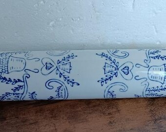May include: A rolling pin with a white ceramic barrel featuring a blue floral design. The handles are made of dark brown wood. The rolling pin is resting on a wooden surface.