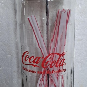 Puede incluir: Un dispensador de pajitas de cristal transparente Coca-Cola con tapa de cromo pulido y varilla metálica central. El cristal presenta el logotipo rojo de Coca-Cola y el texto Delicious and Refreshing. Dentro se ven pajitas blancas con rayas rojas.