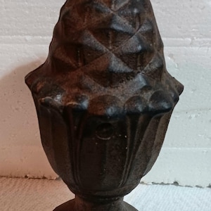Op de afbeelding: Een decoratieve finial van gietijzer met een getextureerde, donkerbruine afwerking. De finial is gevormd als een dennenappel bovenop een gegroefde urn-achtige basis, geschikt voor tuin- of architectuurdecoratie.