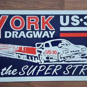 Może przedstawiać: Prostokątny metalowy znak z granatowym tłem zawiera tekst "YORK DRAGWAY" w kolorze czerwonym i białym. Biała grafika samochodu wyścigowego z "US-30" i "the SUPER STRIP" w kolorze białym.