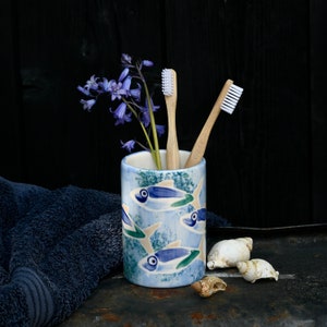 May include: A cylindrical ceramic toothbrush holder decorated with blue and white fish, containing two bamboo toothbrushes and bluebell flowers. A blue towel and seashells are next to the holder. The background is dark.