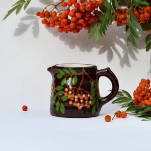 May include: A dark brown ceramic creamer jug featuring a hand-painted design of green leaves and clusters of orange berries. The jug has a curved handle and a small pouring spout, positioned near fresh sprigs of orange rowan berries.