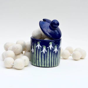 May include: A small blue ceramic jar with a lid, decorated with white snowdrop flowers and green leaves. The jar is filled with white felt balls.